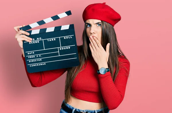 Young brunette teenager holding video film clapboard covering mouth with hand, shocked and afraid for mistake. surprised expression