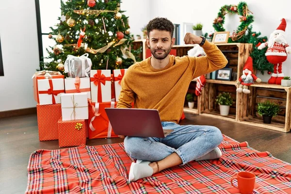 阿拉伯年轻人坐在圣诞树旁的笔记本电脑上 强壮有力地展示着臂膀肌肉 自信而自豪 — 图库照片