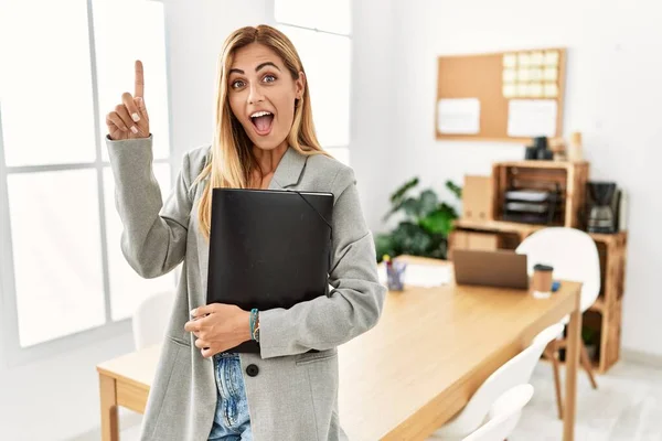 Blonde business woman at the office smiling amazed and surprised and pointing up with fingers and raised arms.