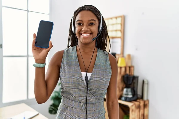 Young african american woman working at the office wearing operator headset holding smartphone looking positive and happy standing and smiling with a confident smile showing teeth