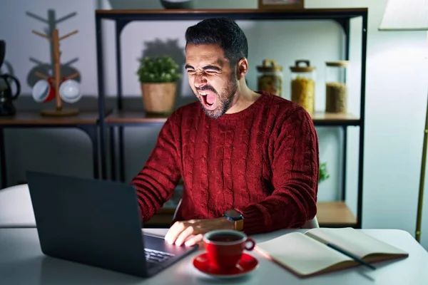 Young hispanic man with beard using computer laptop at night at home angry and mad screaming frustrated and furious, shouting with anger. rage and aggressive concept.