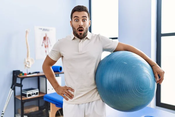 Bonito Homem Hispânico Segurando Bola Pilates Clínica Reabilitação Assustado Surpreso — Fotografia de Stock