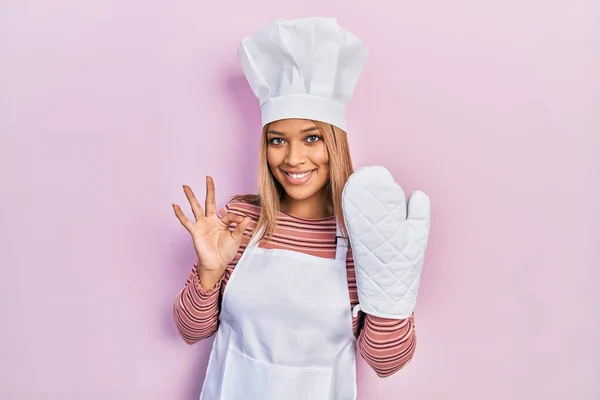 Beautiful hispanic woman uniform wearing apron and hat wearing protective glove doing ok sign with fingers, smiling friendly gesturing excellent symbol