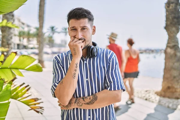Young handsome man listening to music using headphones outdoors looking stressed and nervous with hands on mouth biting nails. anxiety problem.