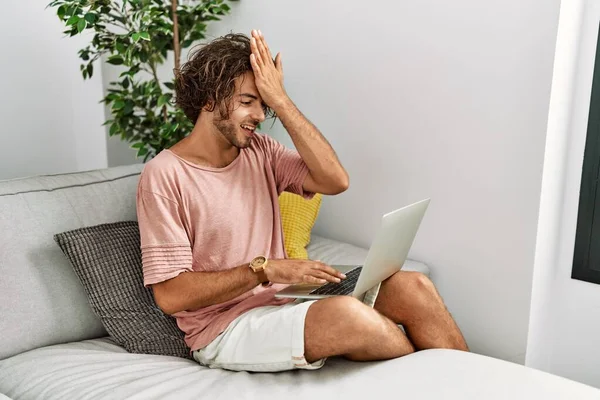 Young hispanic man sitting on the sofa at home using laptop surprised with hand on head for mistake, remember error. forgot, bad memory concept.