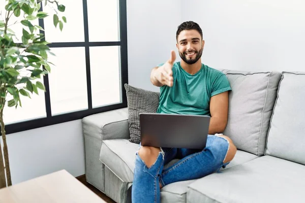 年轻英俊的男人留着胡子 用电脑笔记本电脑坐在沙发上 微笑友好地握手致意和欢迎 成功的商业 — 图库照片