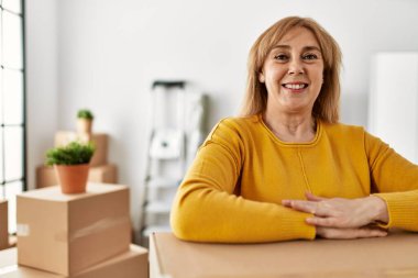 Orta yaşlı sarışın kadın mutlu bir şekilde gülümsüyor. Yeni evindeki karton kutuya yaslanıyor..