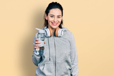 Young hispanic woman wearing sportswear holding water bottle using headphones looking positive and happy standing and smiling with a confident smile showing teeth 