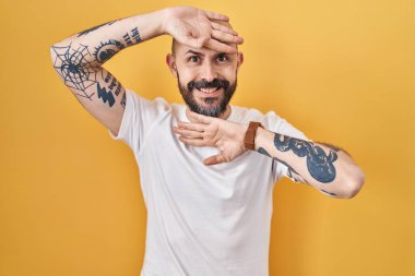 Young hispanic man with tattoos standing over yellow background smiling cheerful playing peek a boo with hands showing face. surprised and exited 