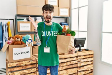Hispanic man with beard wearing volunteer t shirt at donations point celebrating victory with happy smile and winner expression with raised hands 