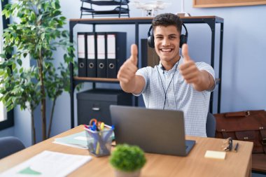 Young hispanic man working at the office wearing headphones approving doing positive gesture with hand, thumbs up smiling and happy for success. winner gesture. 