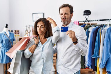 Hispanic middle age couple holding shopping bags and credit card confuse and wondering about question. uncertain with doubt, thinking with hand on head. pensive concept. 