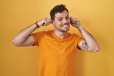 Sarı arka planda dikilen İspanyol genç adam gülümsüyor parmaklarıyla kulaklarını çekiyor, komik bir jest. deneme sorunu 