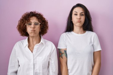 Hispanic mother and daughter wearing casual white t shirt over pink background puffing cheeks with funny face. mouth inflated with air, crazy expression. 
