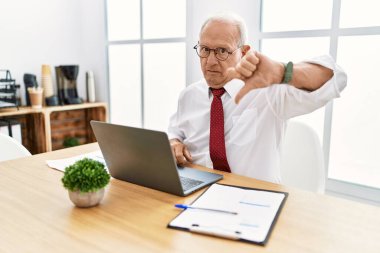 Senior man working at the office using computer laptop looking unhappy and angry showing rejection and negative with thumbs down gesture. bad expression. 