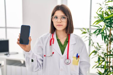 Young hispanic doctor woman showing smartphone screen thinking attitude and sober expression looking self confident 