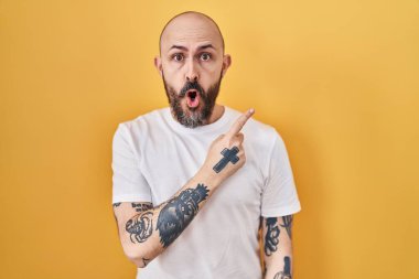 Young hispanic man with tattoos standing over yellow background surprised pointing with finger to the side, open mouth amazed expression. 