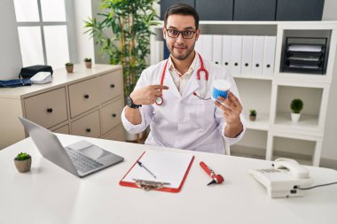Young hispanic doctor man with beard holding ear cotton buds pointing finger to one self smiling happy and proud 