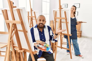 Senior artist man at art studio smiling cheerful presenting and pointing with palm of hand looking at the camera. 