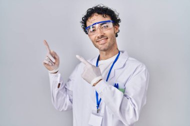 Hispanic man working at scientist laboratory smiling and looking at the camera pointing with two hands and fingers to the side. 