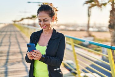 Middle age hispanic woman working out with smartphone at promenade