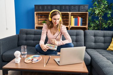 Young beautiful woman practicing painting looking at online tutorial thinking attitude and sober expression looking self confident 