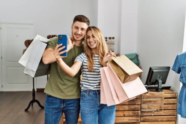 Young hispanic customer couple holding shopping bags and make selfie by the smartphone at clothing store.