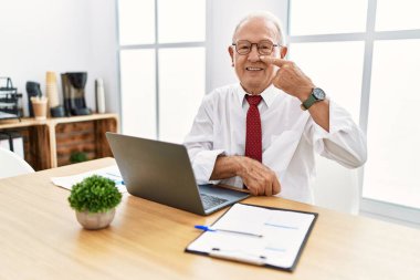 Senior man working at the office using computer laptop pointing with hand finger to face and nose, smiling cheerful. beauty concept 