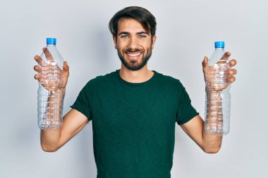 Young hispanic man holding recycling plastic bottles smiling with a happy and cool smile on face. showing teeth. 