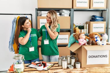 Two young women wearing volunteer t shirt at donations stand serious face thinking about question with hand on chin, thoughtful about confusing idea 