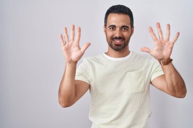 Sakallı İspanyol adam izole edilmiş arka planda duruyor ve parmaklarını on numarayı gösterip işaret ederken kendinden emin ve mutlu bir şekilde gülümsüyor.. 
