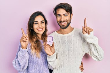 Beautiful couple of boyfriend and girlfriend holding engagement ring for proposal surprised with an idea or question pointing finger with happy face, number one 