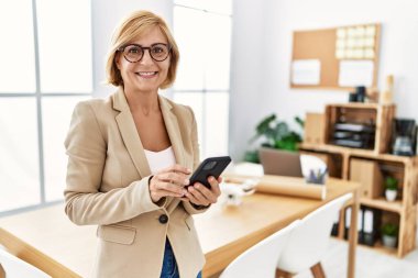 Mimarlık stüdyosunda çalışan orta yaşlı sarışın kadın akıllı telefon kullanıyor.