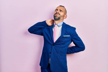 Genç İspanyol bir adam iş elbisesi giyiyor ve kravat takıyor. Boyun ağrısı çekiyor. Boynuna elinle dokunuyor. 