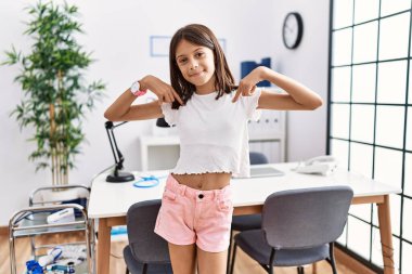 Young hispanic girl standing at pediatrician clinic looking confident with smile on face, pointing oneself with fingers proud and happy. 