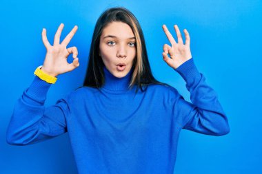 Young brunette girl wearing turtleneck sweater looking surprised and shocked doing ok approval symbol with fingers. crazy expression 