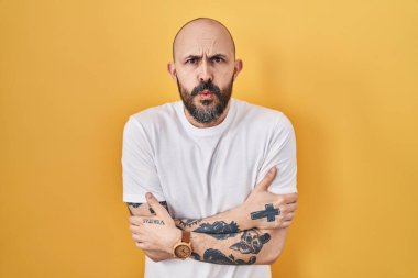 Young hispanic man with tattoos standing over yellow background shaking and freezing for winter cold with sad and shock expression on face 