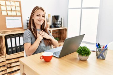 Young brunette woman working at the office with laptop smiling with hands on chest with closed eyes and grateful gesture on face. health concept. 