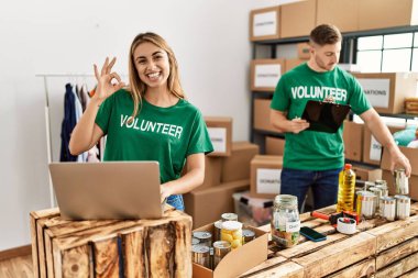 Young woman and man wearing volunteer t shirt at donations stand doing ok sign with fingers, smiling friendly gesturing excellent symbol 