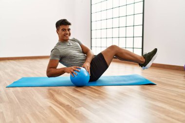 Genç İspanyol adam gülümsüyor. Spor merkezinde top kullanarak karın kasları çalıştırıyor.