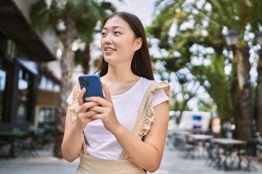 Genç Çinli kız şehirde akıllı telefon kullanarak mutlu bir şekilde gülümsüyor..