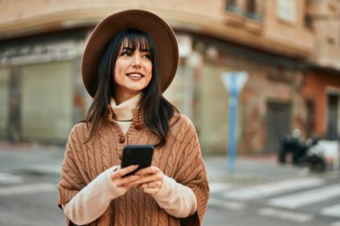 Kışlık şapkalı esmer kadın, şehirde akıllı telefon kullanarak gülümsüyor.