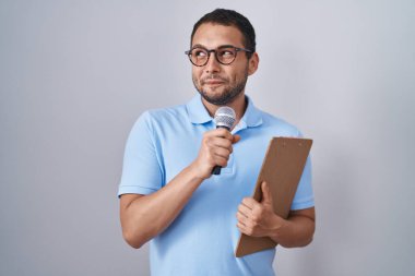 Hispanic man holding reporter microphone and clipboard smiling looking to the side and staring away thinking. 