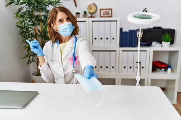 Middle age doctor woman offering safety mask smiling happy pointing with hand and finger to the side