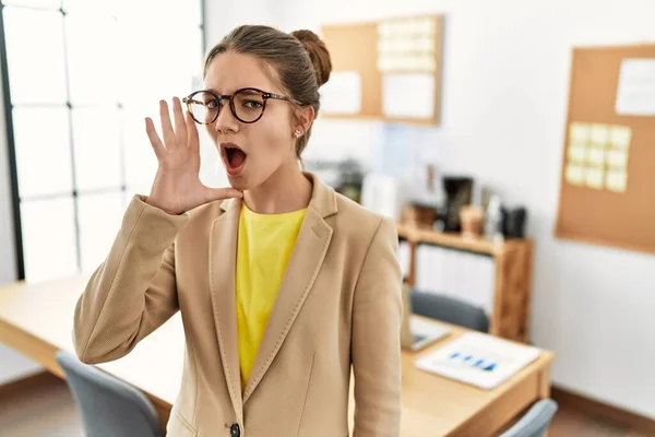 Young brunette teenager wearing business style at office shouting and screaming loud to side with hand on mouth. communication concept.