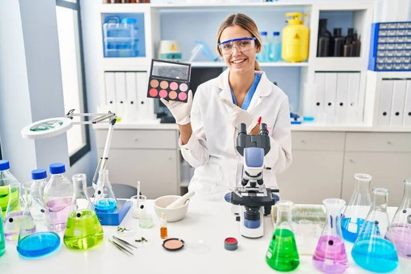 Young blonde woman working at scientist laboratory with make up smiling happy pointing with hand and finger