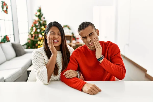 一对年轻的拉丁夫妇坐在桌子上 坐在圣诞树旁 手牵着手 带着痛苦的表情摸着嘴 因为牙齿疼痛或牙病 牙医概念 — 图库照片