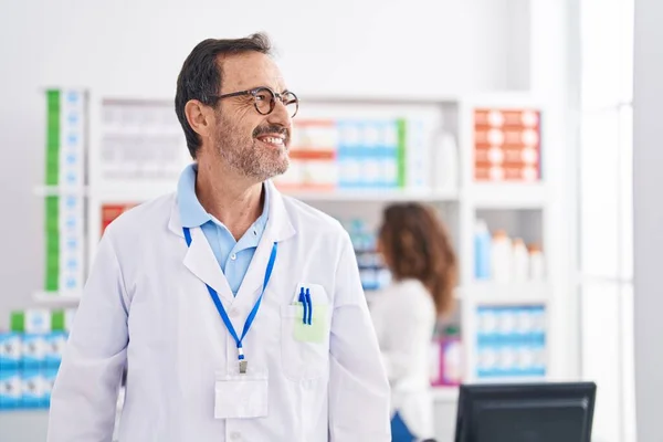 Middle age hispanic man working at pharmacy drugstore looking to side, relax profile pose with natural face and confident smile.