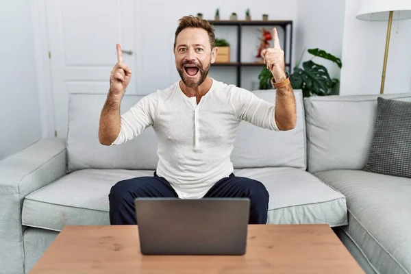 Middle age man using laptop at home smiling amazed and surprised and pointing up with fingers and raised arms.
