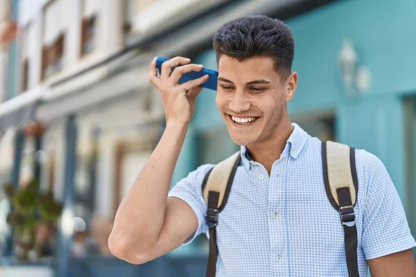 Young hispanic man student listening audio message by smartphone at street